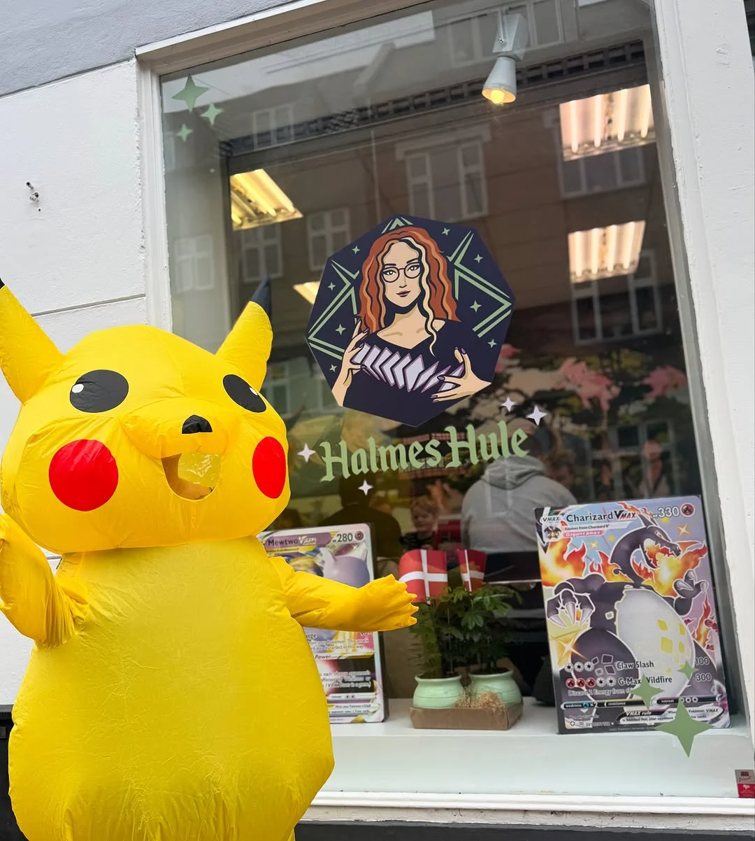 En stor Pikachu-figur foran Halmes Hules butiksvindue med Pokémon-kort og butikkens logo.